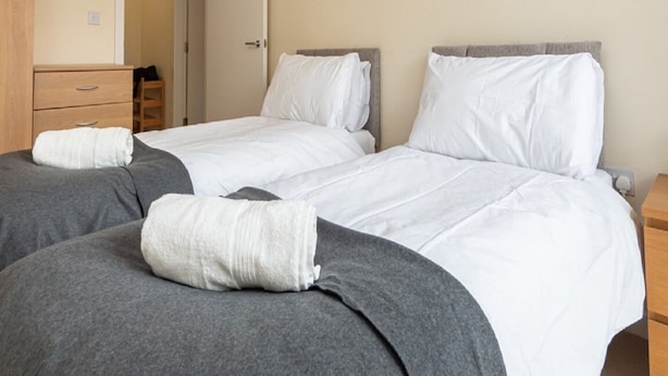Ein Hotelzimmer mit zwei Einzelbetten, jeweils mit einer grauen Decke und weißen Kissen. Es gibt einen Holzschrank und einen kleinen Nachttisch mit einer Lampe.