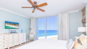 Un dormitorio con una cama grande, un ventilador de techo, un aparador, una silla y un televisor mostrando una escena de playa.