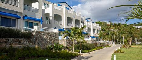 A row of white buildings with blue accents, a stone wall, and a paved walkway with palm trees.