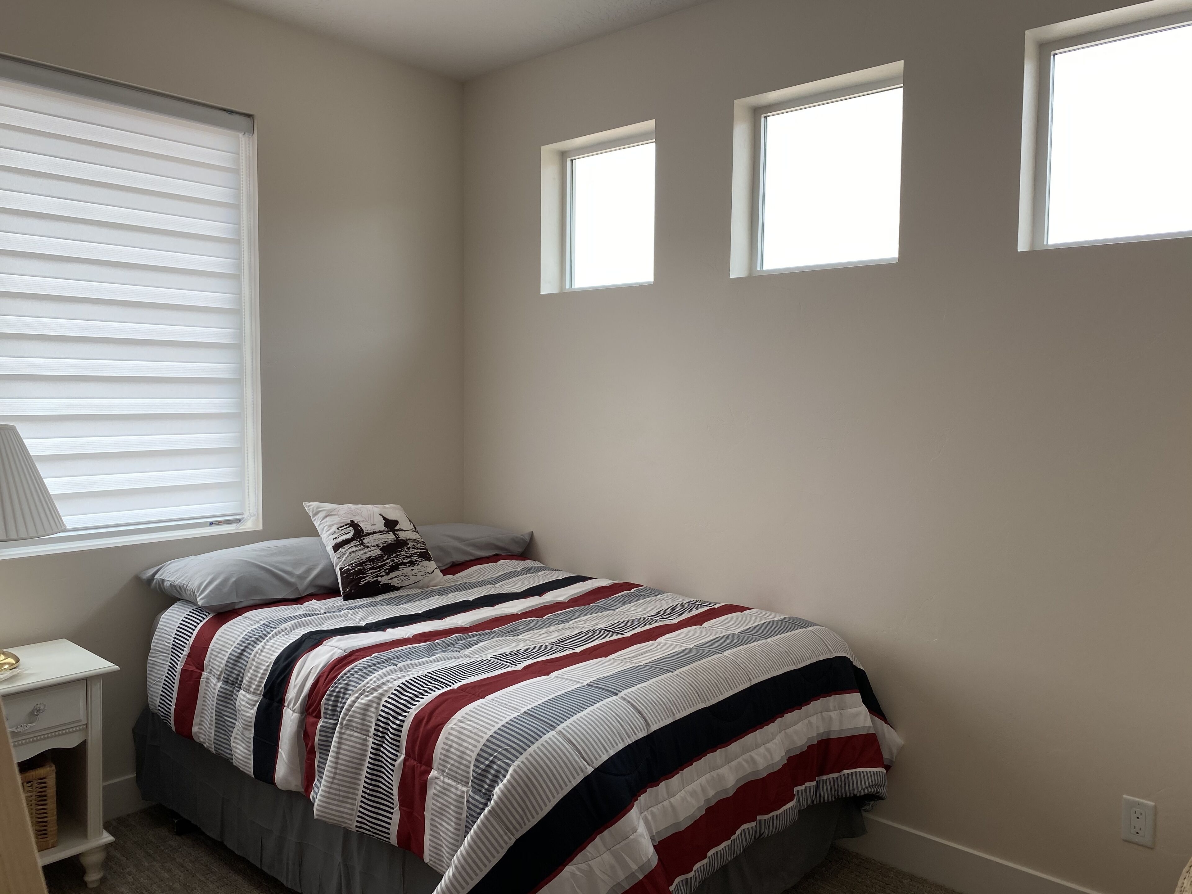 Un dormitorio con una cama, una mesita de noche, una lámpara y tres ventanas.