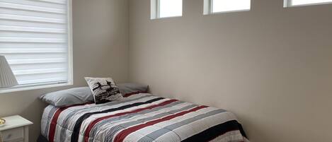Un dormitorio con una cama, una mesita de noche, una lámpara y tres ventanas.
