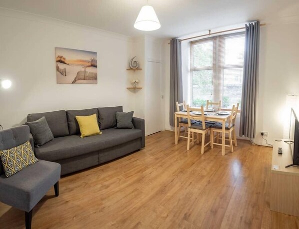 Een woonkamer met een grijze bank, een eettafel met stoelen, een schilderij aan de muur en een groot raam met gordijnen.
