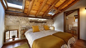 A bedroom with a stone wall, wooden beams, a skylight, a bed with two pillows, and a bathroom visible through an open door. - El Pajar de Justa (Adrados)