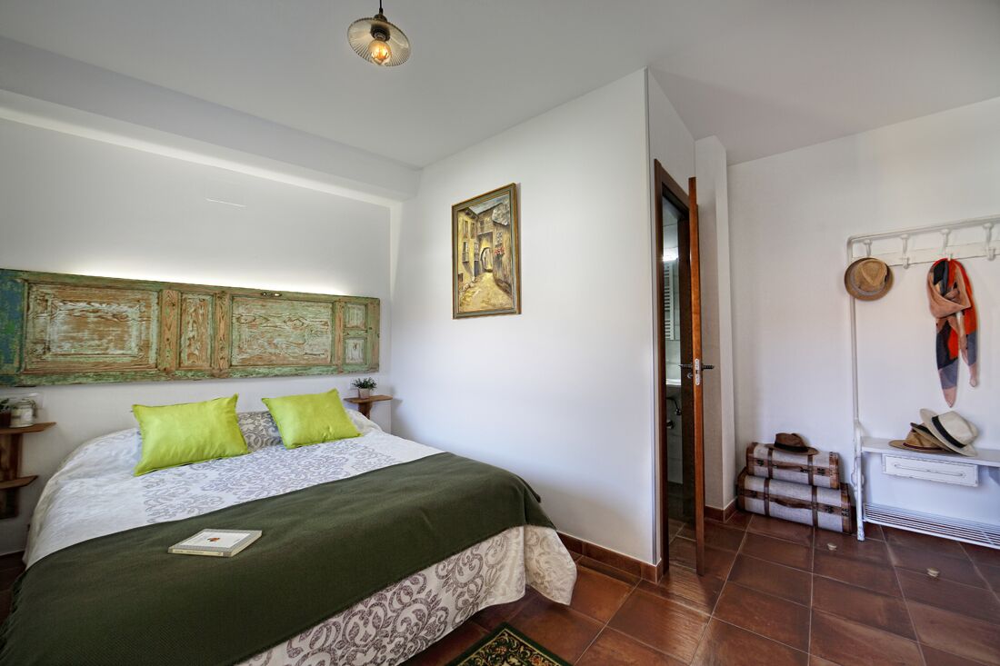 Een slaapkamer met een bed, een schilderij, een hoedenrek en een klein tafeltje.