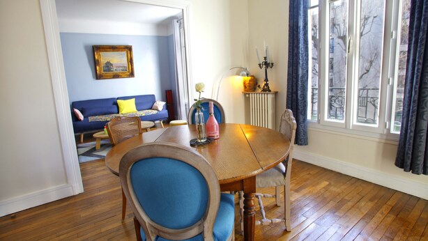 Un comedor con una mesa y sillas de madera, un sofá azul al fondo y una ventana con cortinas.