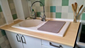 Una cocina con un fregadero, un grifo, un jabonera, una botella de detergente, una tabla de cortar, utensilios de madera y un microondas.