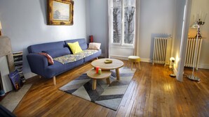 Una sala de estar con un sofá azul, una pequeña mesa redonda, un radiador y una ventana con vistas a árboles.