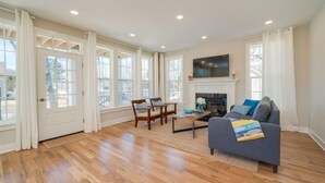 A spacious living room with hardwood floors, a fireplace, a television mounted above it, and large windows with curtains. - Beach Breeze - 5 bedroom 4.5 bath vacation rental located just a block from the beach! (South Haven)