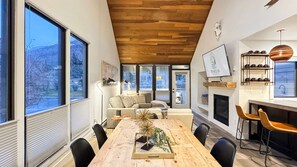 A modern interior with a wooden ceiling, large windows, a dining area with a wooden table, and a fireplace.