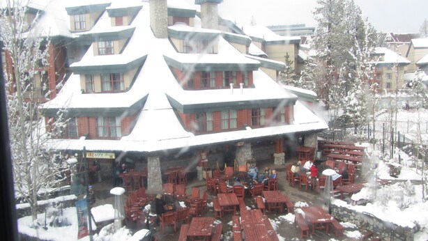 A building with a snow-covered roof and multiple windows. There are outdoor tables and chairs arranged in front of the building.