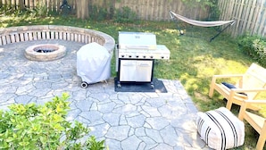 Un jardin avec une terrasse en pierre, un barbecue couvert, un foyer, un brasero, un hamac et une chaise en bois.