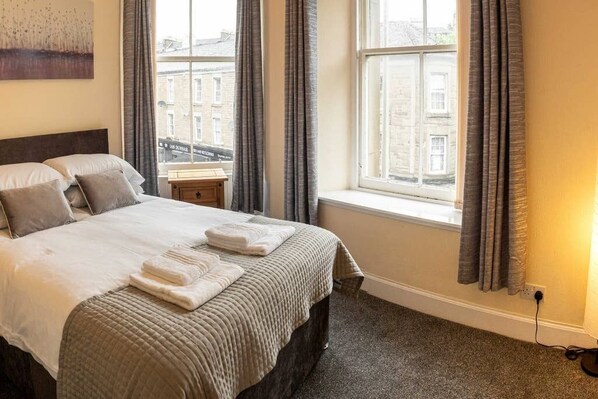 A hotel room with a large bed, a nightstand, a window with curtains, and a view of buildings outside.