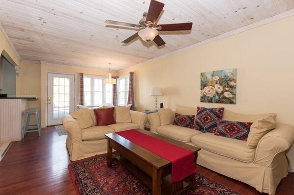 Un salon avec un plafond en bois, un ventilateur de plafond, un ensemble de canapés beige, une table basse et un plaid rouge.