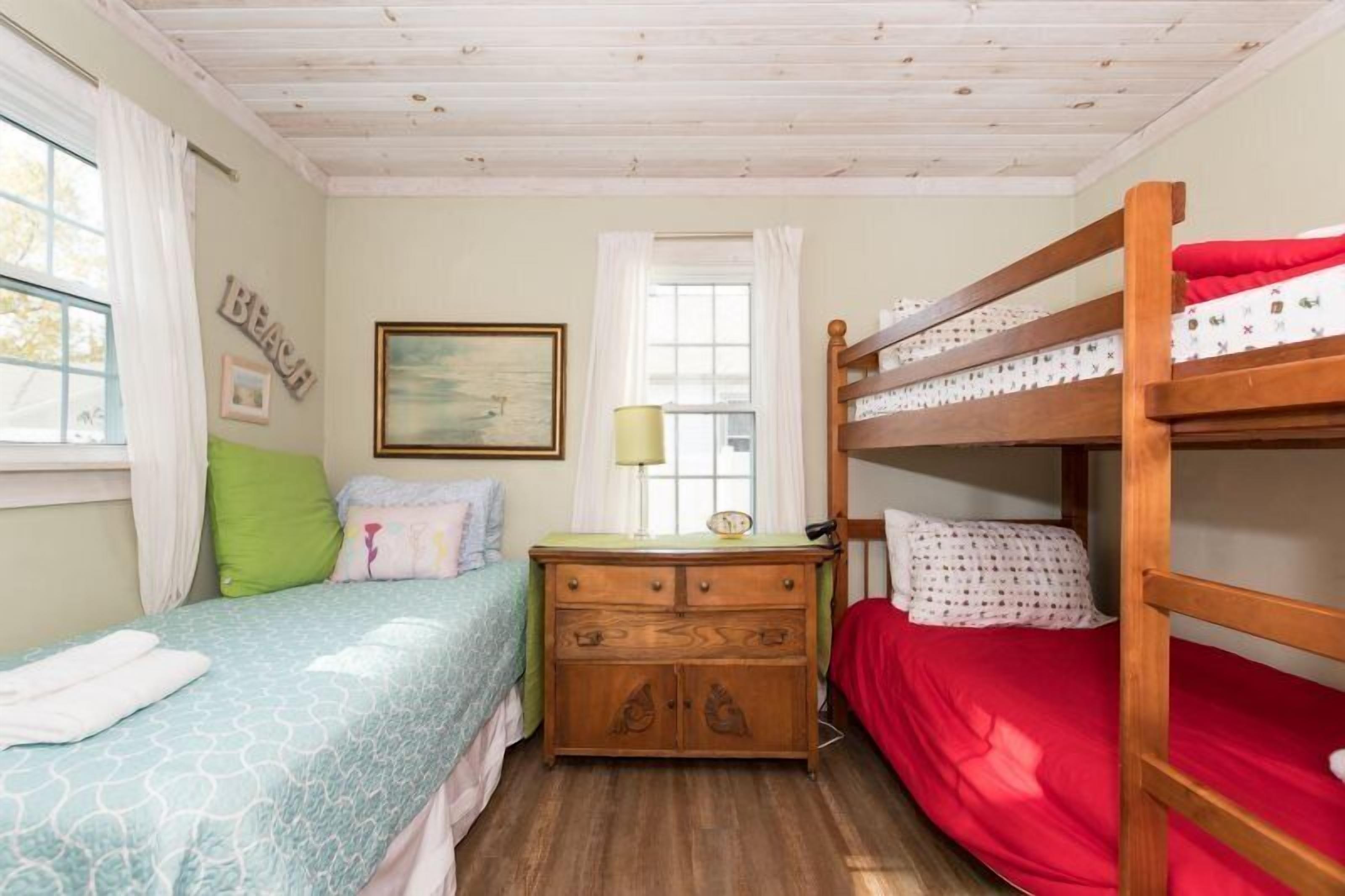 A room with two beds, one with a blue patterned bedspread and the other with a red bedspread. There is a wooden dresser with a lamp and a framed picture on the wall.