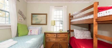 A room with two beds, one with a blue patterned bedspread and the other with a red bedspread. There is a wooden dresser with a lamp and a framed picture on the wall.