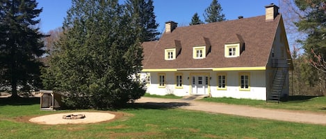 Une maison à deux étages, avec un toit en pente, une cheminée et un jardin devant, où se trouve un foyer.