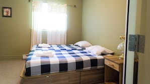 A single bed with a checkered bedspread, a bedside table with a lamp, and a window with curtains. - Villa st-Léon (Saint-Gabriel-de-Valcartier)