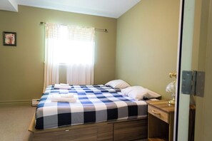 A single bed with a checkered bedspread, a bedside table with a lamp, and a window with curtains. - Villa st-Léon (Saint-Gabriel-de-Valcartier)