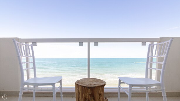 Un balcon avec deux chaises blanches et une petite table en bois, donnant sur une plage aux eaux calmes.