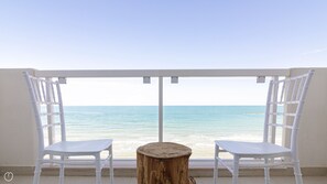 A balcony with two white chairs and a small wooden table, overlooking a beach with calm waters.