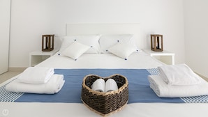 A neatly made bed with white linens, a blue bedspread, and a wicker basket in the center containing two white towels.