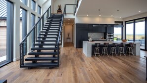 Eine moderne Küche mit Kochinsel und Barhocker, eine Treppe mit Glasgeländern und große Fenster mit Blick ins Freie.