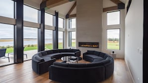 Ein modernes Wohnzimmer mit einer großen, winkelförmigen Couchgarnitur, einem runden Couchtisch und einem flachbildfernseher an der Wand. Große Fenster bieten Blick ins Freie.
