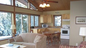 Una sala de estar acogedora con un sofá, una mesa de centro y una mesa de comedor. Hay una zona de cocina con armarios de madera, un microondas y una estufa. Ventanas amplias ofrecen luz natural y vistas a árboles.