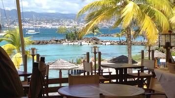A seaside restaurant with wooden tables and chairs, a view of the ocean, and palm trees.