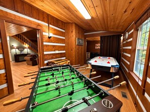 A room with a foosball table, a pool table, wooden walls, and a staircase leading to another area.