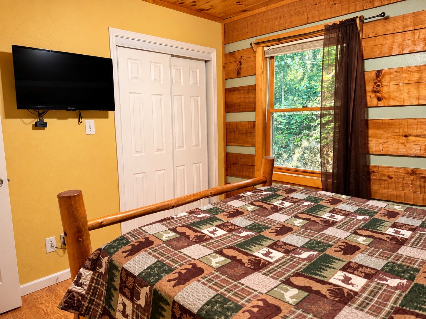 A room with a bed, a television mounted on the wall, a window with curtains, and a door.