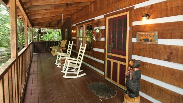 Een houten hutje met een veranda, schommelstoelen, een beerbeeld en een welkomstbord.