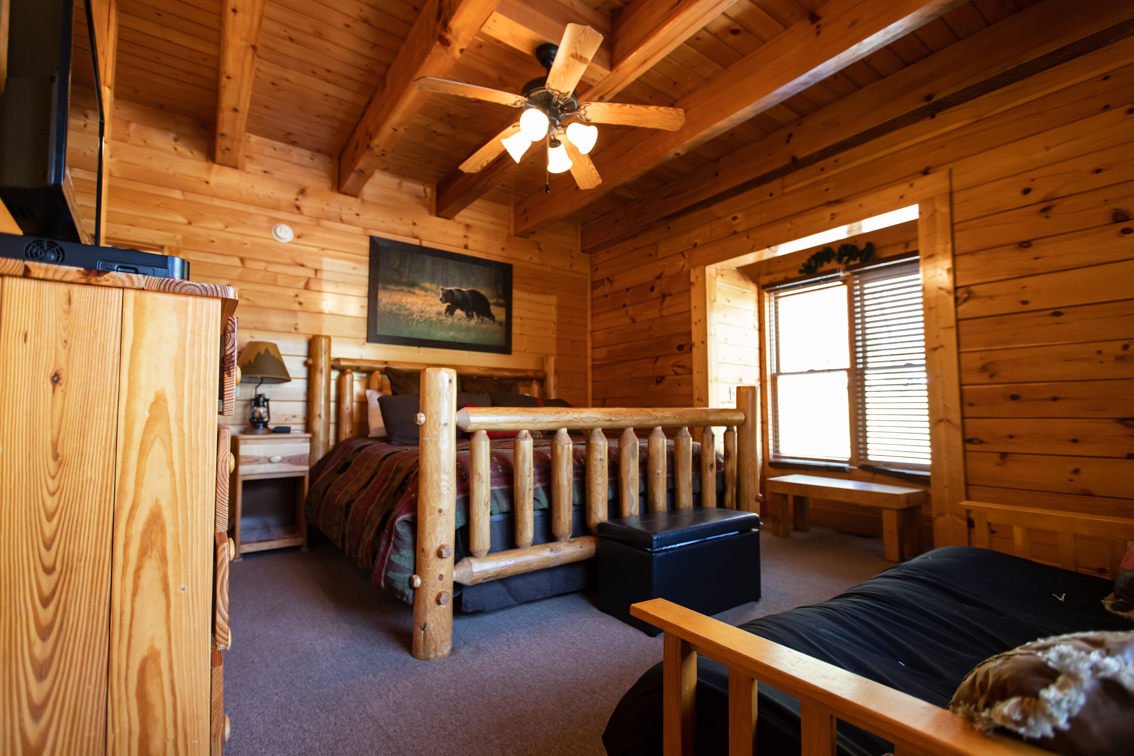 A log cabin room with a bed, a desk, a chair, a ceiling fan, and a window with blinds.
