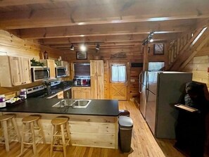 Uma cozinha compacta com armários de madeira, uma ilha central com banquetas, micro-ondas, geladeira e uma janela com cortinas.