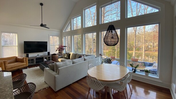 A spacious living room with a large window, a flat-screen TV, a sofa, a dining table, and a chandelier. - Pond View Retreat: Your Ultimate Athens Getaway Minutes To Sanford Stadium (Athens)