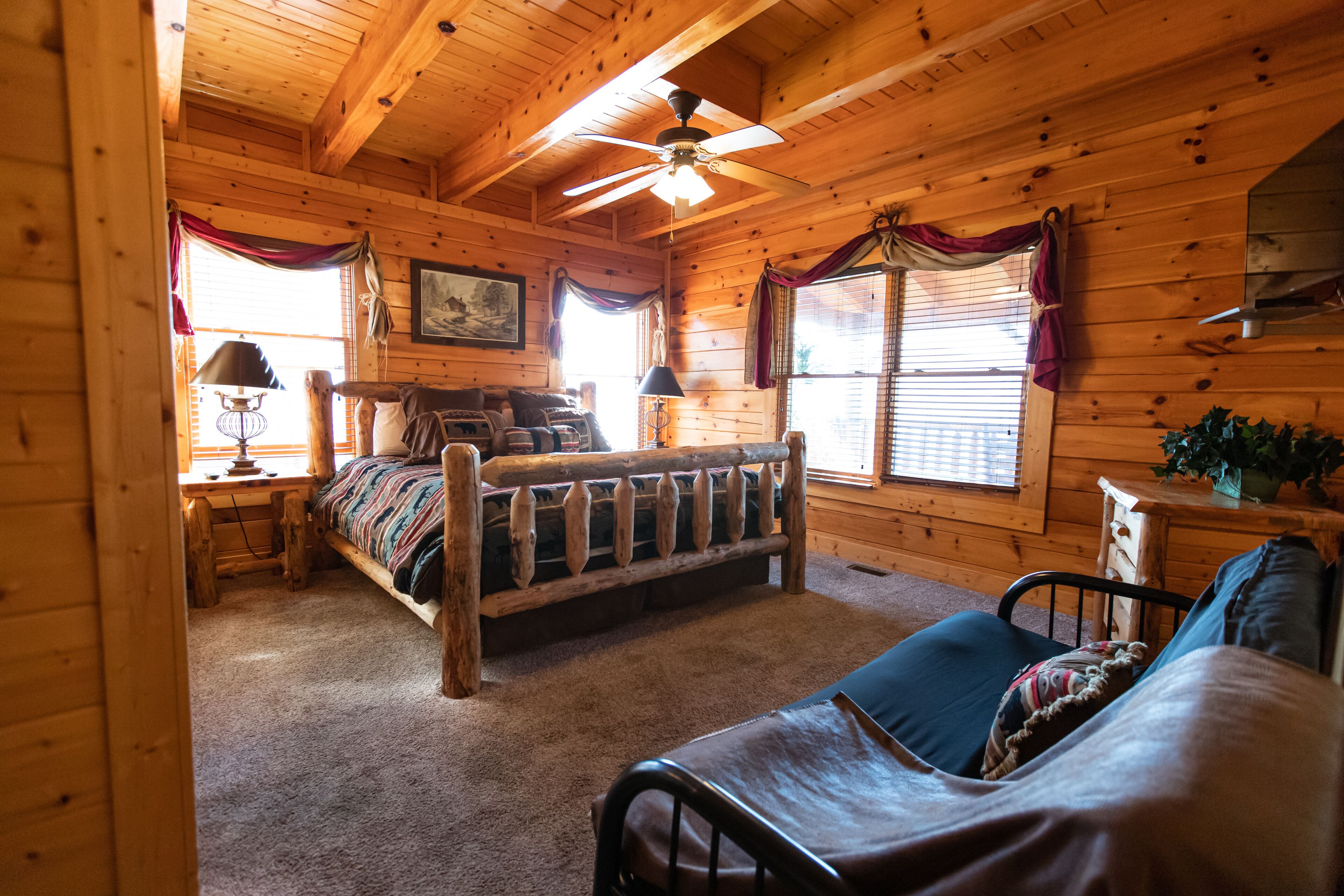 A cozy cabin-style room with a wooden ceiling, a bed with a headboard, a desk with a lamp, and a window with curtains.