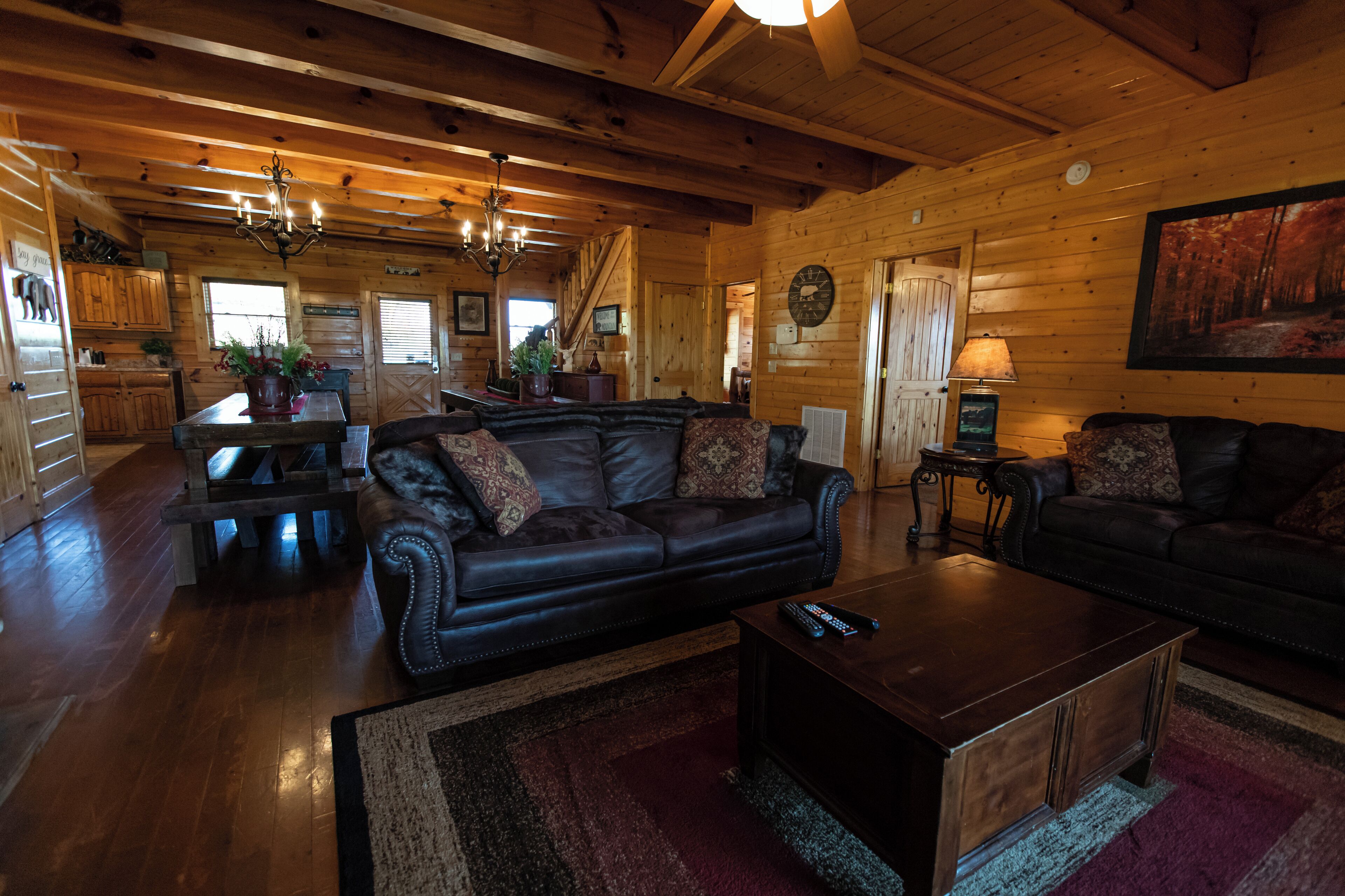 A spacious living room with a large black leather sofa, a wooden coffee table, and a chandelier. A kitchen area with wooden cabinets and a dining table is visible in the background.