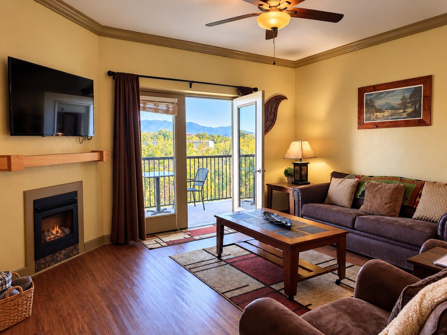 A living room with a fireplace, a television, a sofa, a coffee table, and a balcony with a view of trees and mountains.