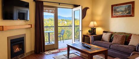 A living room with a fireplace, a television, a sofa, a coffee table, and a balcony with a view of trees and mountains.