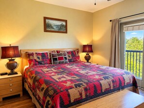 A bedroom with a large bed, two bedside lamps, a nightstand, a window with curtains, and a painting on the wall.