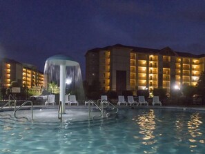 Una piscina con una fuente, sillas de descanso y un edificio de hotel al fondo.