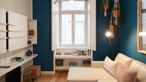 A cozy living room with a white sofa, a small table, and a bookshelf. A window with a white frame allows natural light to enter. - FLH Chiado Nature Apartment (Lisbon)