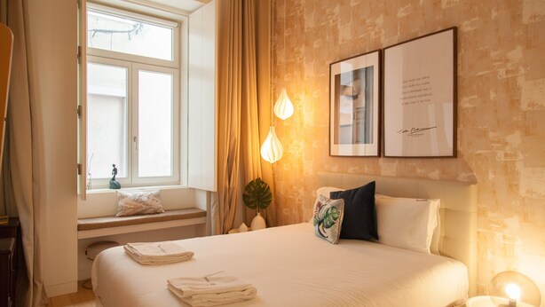 A hotel room with a large bed, a window with curtains, a bedside table with a lamp, and two framed pictures on the wall. - FLH Chiado Nature Apartment (Lisbon)