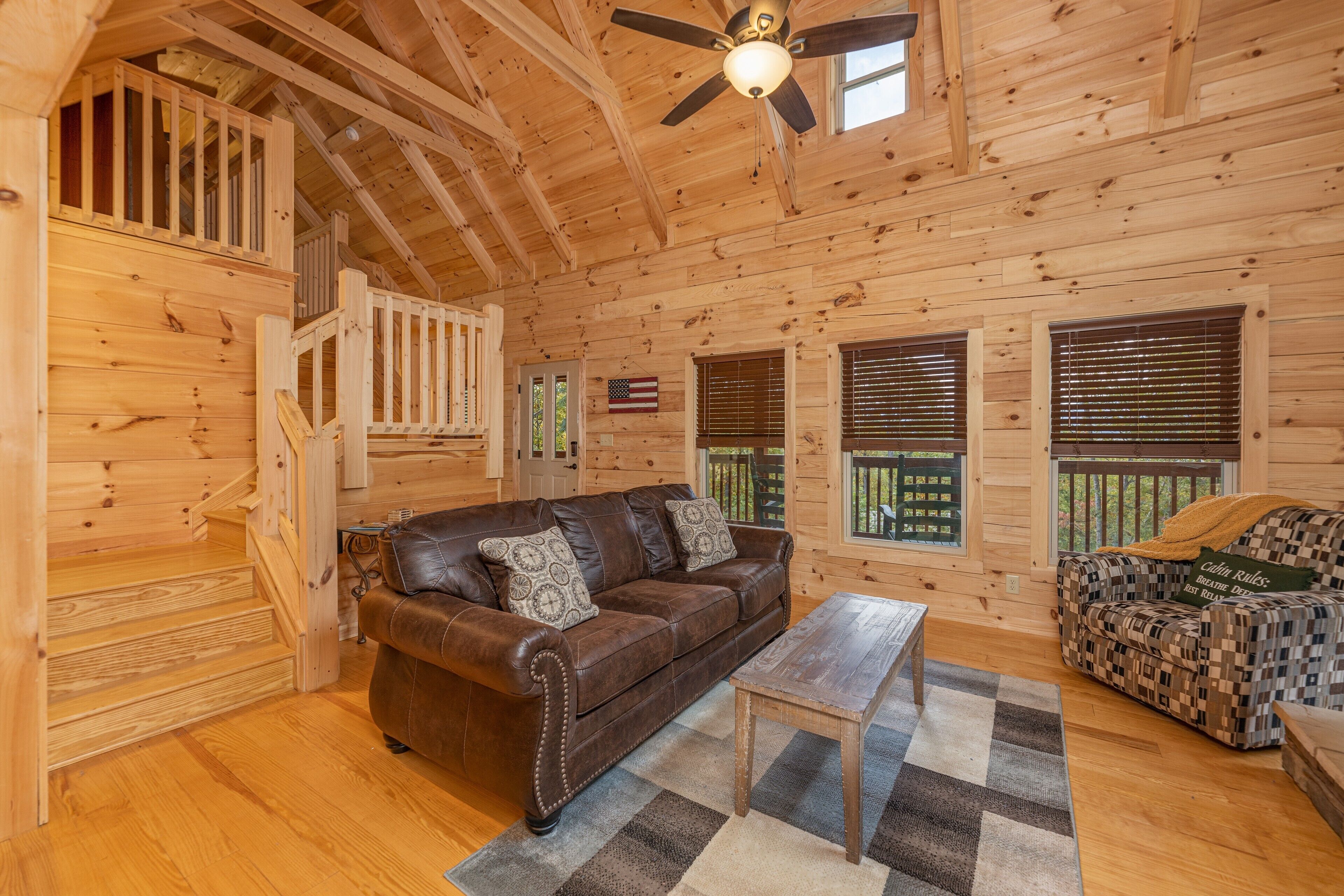 A wooden cabin interior with a brown leather sofa, a patterned sofa, a wooden coffee table, and a staircase leading to an upper level.