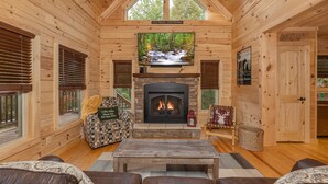 A cozy cabin interior with a stone fireplace, wooden walls, a large TV mounted above it, and a comfortable seating area with a patterned armchair and a couch. - Smoky Retreat (Gatlinburg)