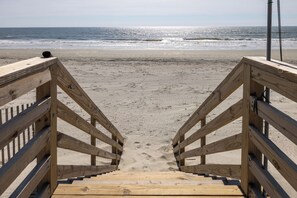 Un escalier en bois mène à une plage de sable, avec l’océan et ses vagues en arrière-plan.