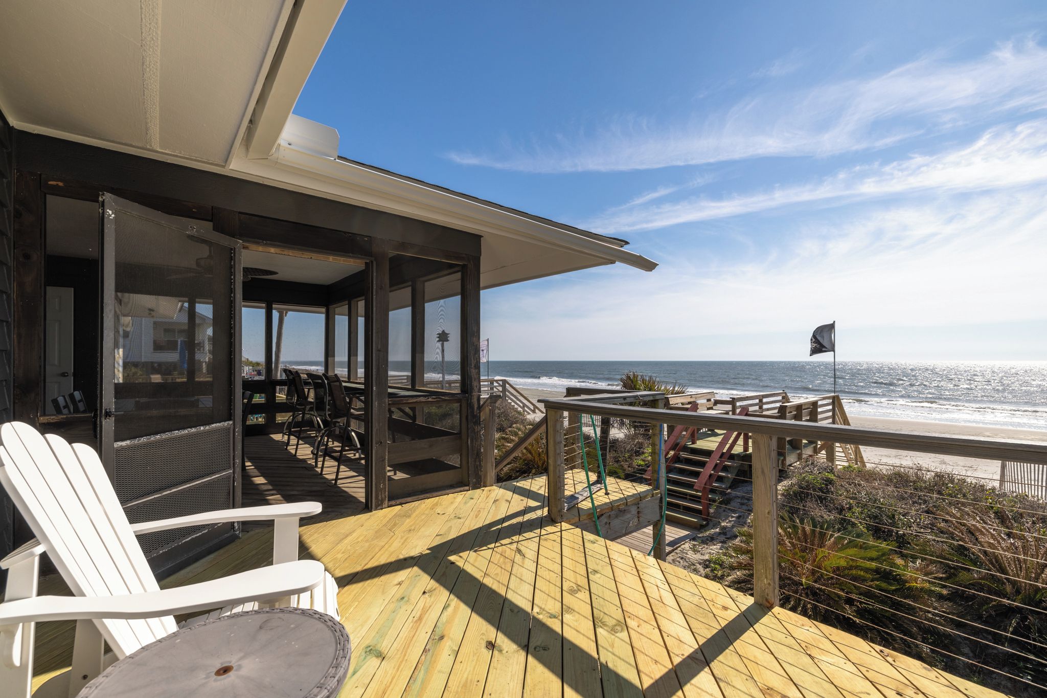 Eine Holzterrasse mit Sitzgelegenheiten im Freien, mit Blick auf einen Strand und das Meer, wo ein Segelboot fährt.