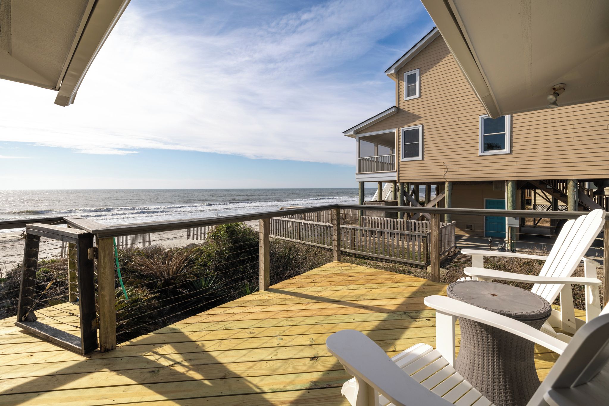 A wooden deck with a view of the ocean, a beach, and a two-story building with a balcony.