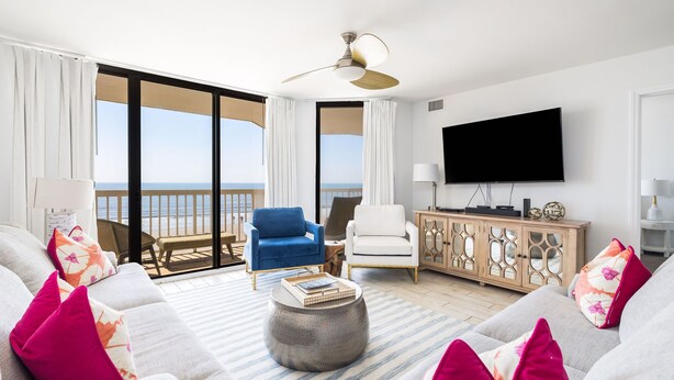 Un salon moderne doté d’un grand téléviseur, de sièges confortables, d’un balcon avec vue sur la plage et d’un ventilateur de plafond.