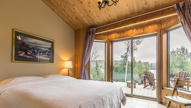 A bedroom with a large bed, a painting, a lamp, and a view of the outdoors through a sliding glass door. - Welcome to the Chalets du Lac Grenier (Chertsey)